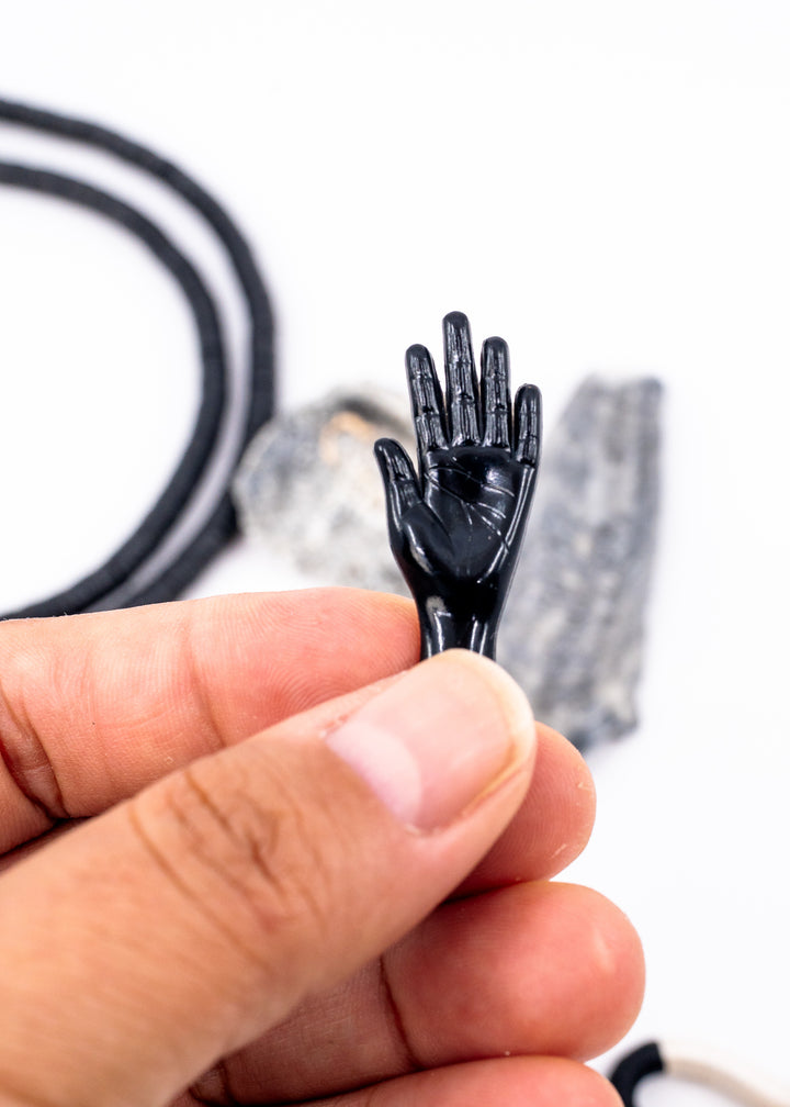 A close-up of a hand holding a small glossy black hand-shaped charm, with blurred black cords and gray rectangular pieces in the background on a white surface. Made from German resin.