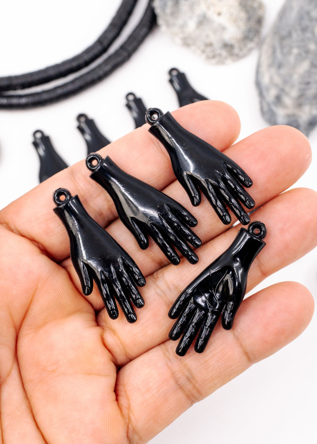 Five glossy black hand-shaped jewelry charms or pendants displayed on an open hand, with additional similar pieces blurred in the background. Made from German resin.