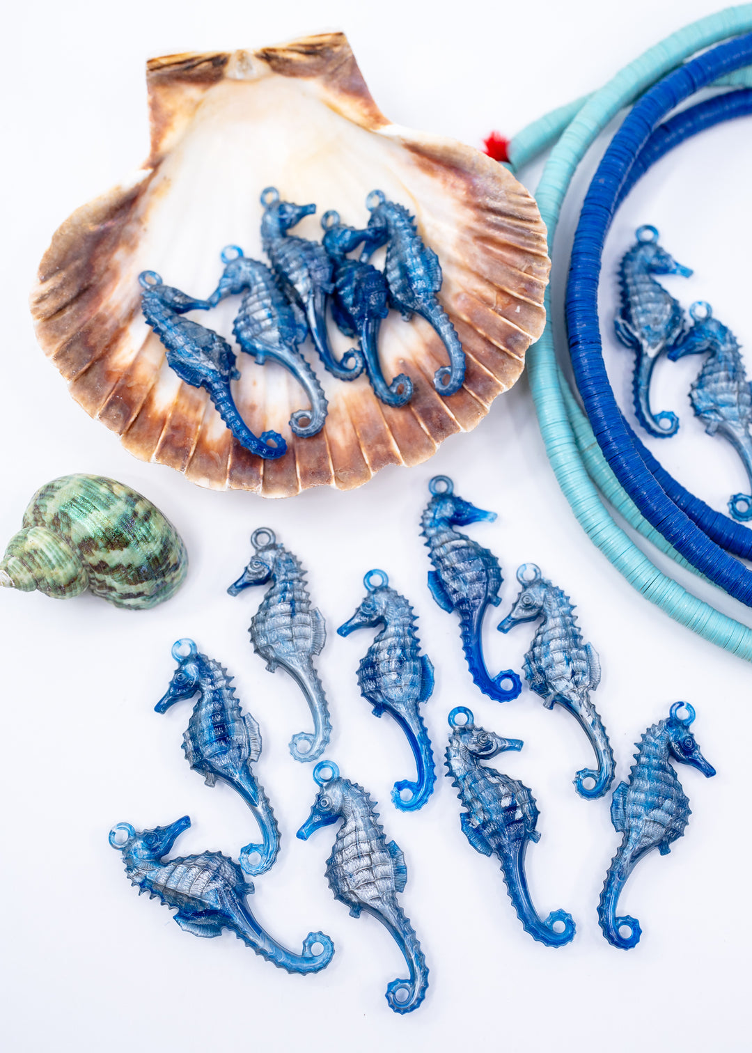 Blue seahorse-shaped jewelry components or charms arranged on a white background, with a scallop shell, a small green seashell, and blue wrapped cords used as styling props. Made from German resin.