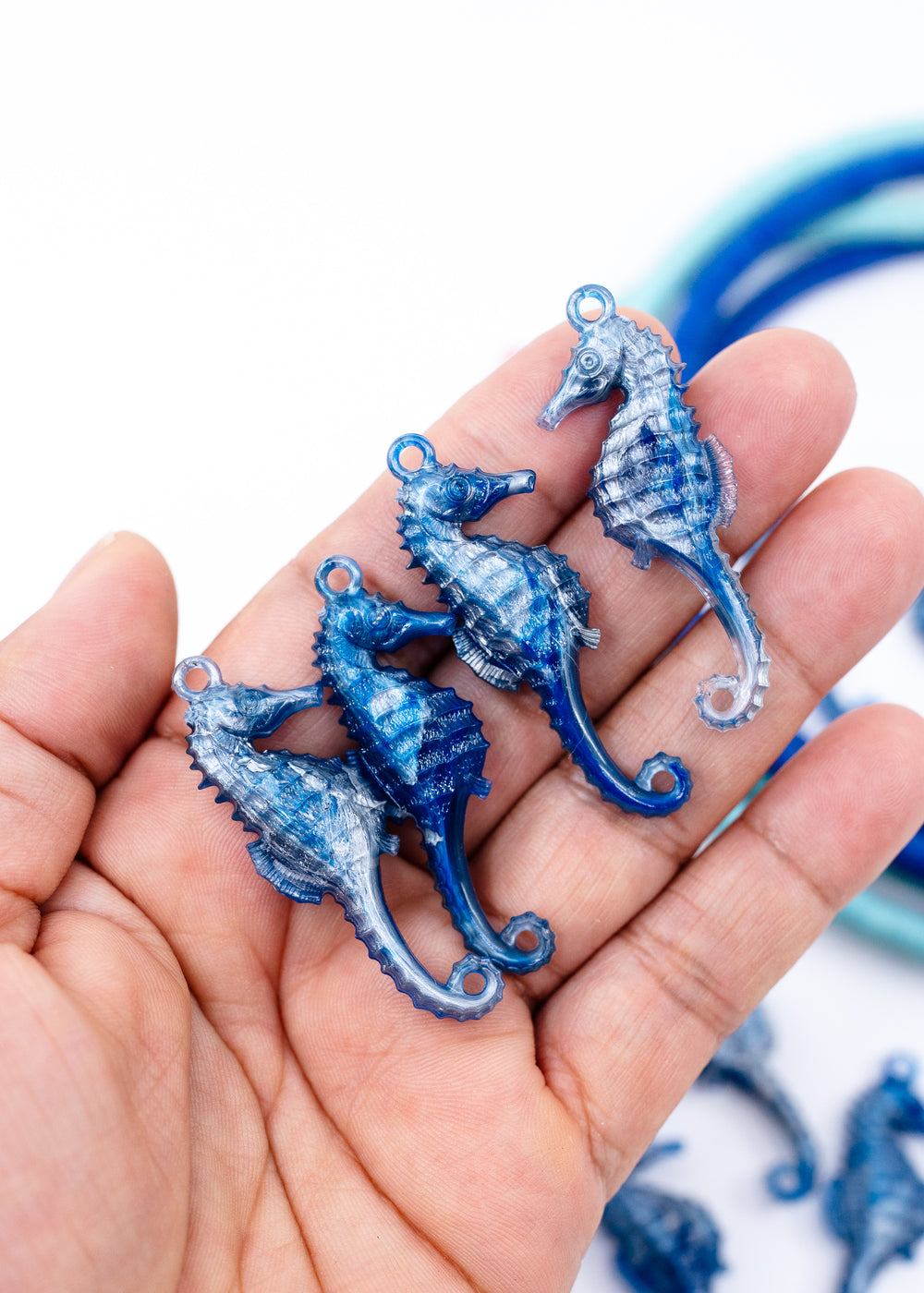 Three blue seahorse-shaped jewelry charms held in a person's hand against a white background. Made from German resin.