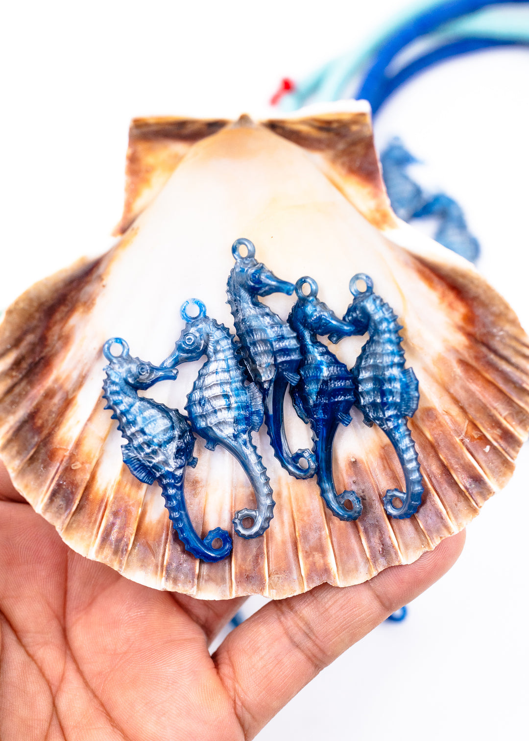 Four blue seahorse charms with silver-tone loops, displayed on a brown seashell held in a person&