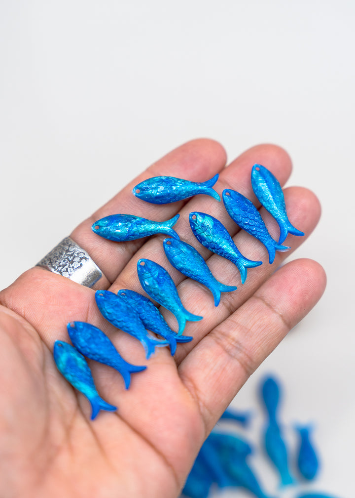 A hand holding several small glittery blue fish-shaped charms or beads against a plain light background.