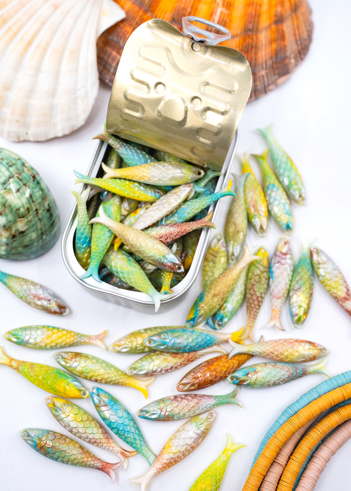 A tin of small decorative fish with many matching sardine fish scattered around it on a white surface, styled with seashells and colorful curved accents. Made in Germany from resin for Jewelry making.