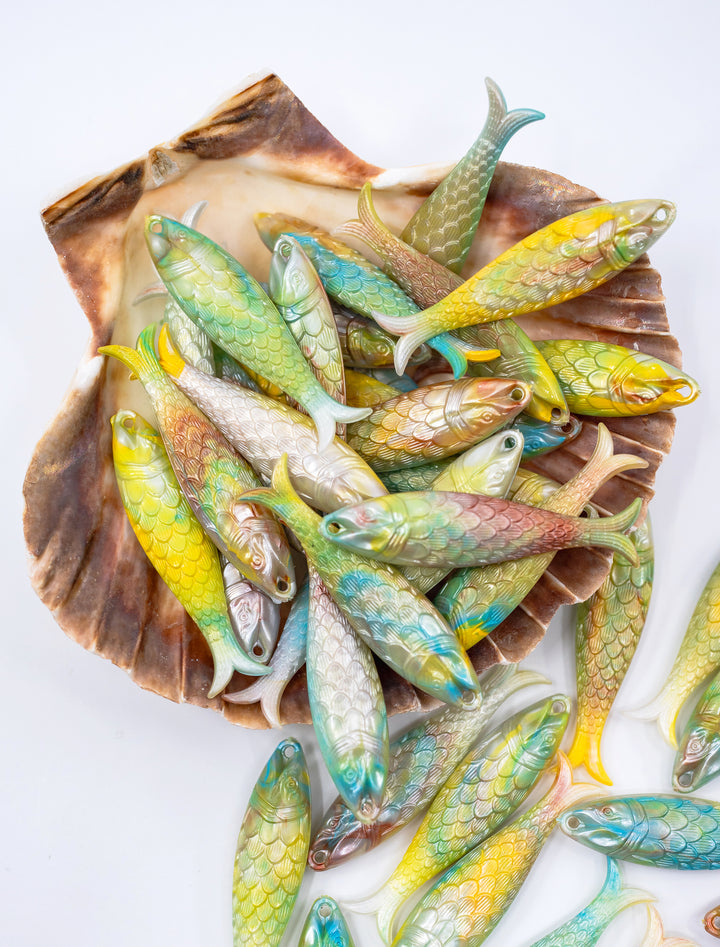 A collection of small, decorative fish-shaped objects with iridescent green, blue, yellow, and pink scales, arranged inside and around a large brown shell on a white background.