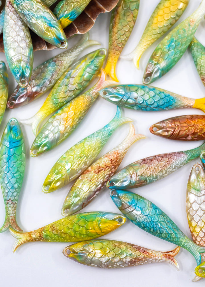 A pile of small decorative fish-shaped items with detailed scales, shown in assorted colors including blue, green, yellow, orange, and brown on a white background. Made in German from German resin.