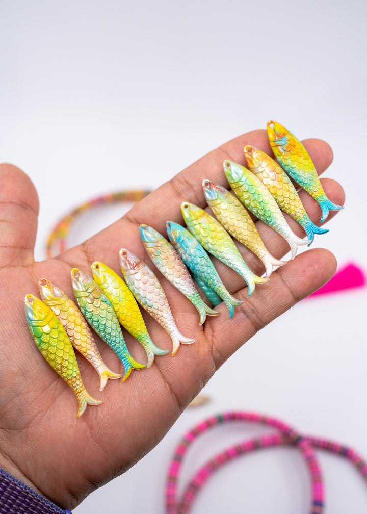 A hand holds a row of small, colorful, fish-shaped charms with detailed scales. The charms feature a gradient of yellow, green, blue, and pink, and are displayed against a clean white background with blurred accessories in the foreground. made in Germany.