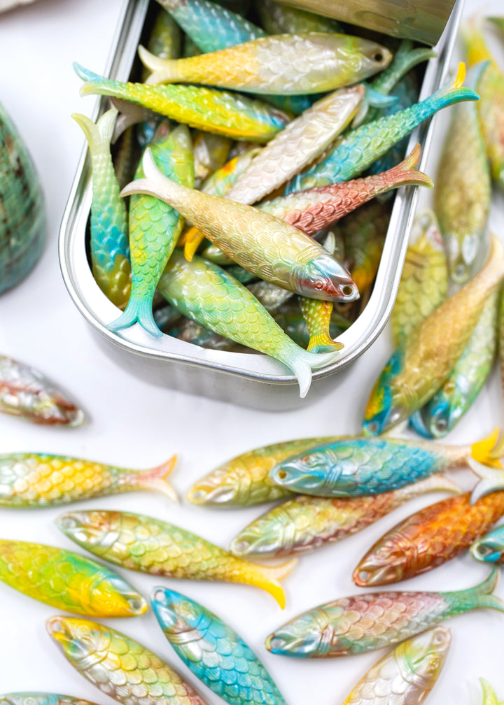 A close-up of many small decorative fish-shaped charms in assorted colors, with several spilled from a rectangular metal container onto a white surface. These fish charms are used in jewelry making.