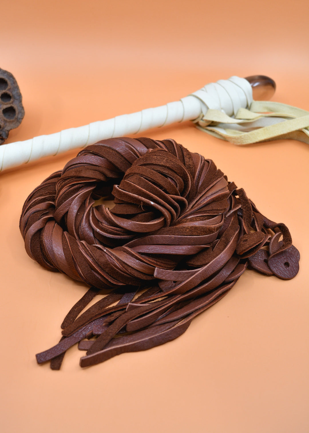 A chocolate brown deerskin leather strap with a smooth texture on one side and a soft suede finish on the other, displayed on an orange background alongside a white object, possibly a tool for crafting.