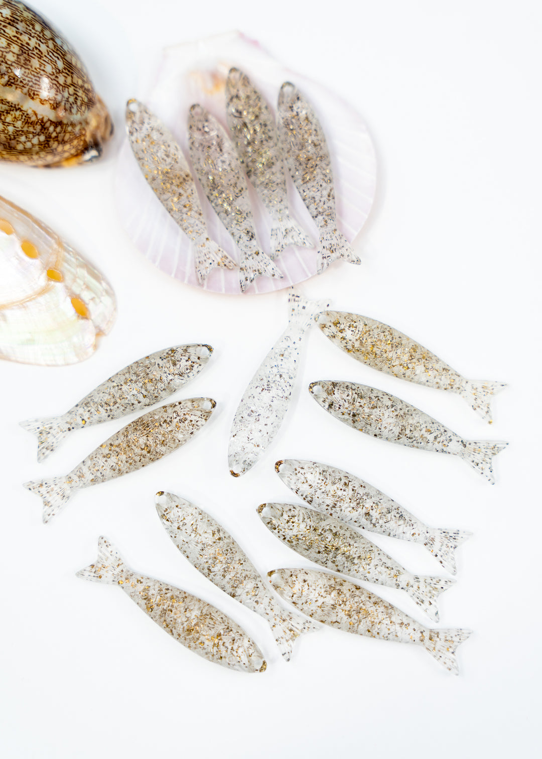 Ten small fish-shaped jewelry charms with a textured, speckled silver and gold finish are arranged on a white background, with decorative seashells in the upper left corner.