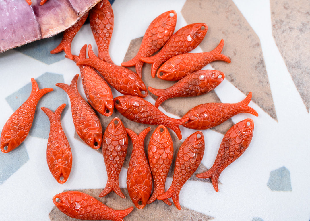 A group of small fish-shaped beads or charms in a glossy terracotta orange-red color, arranged on a light surface with a few pieces partially visible in a purple container at the top left.