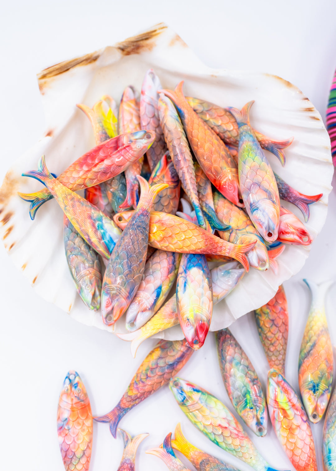 A pile of small decorative fish-shaped charms made from German resin with colorful painted scales, arranged on a white shell and scattered on a white surface.