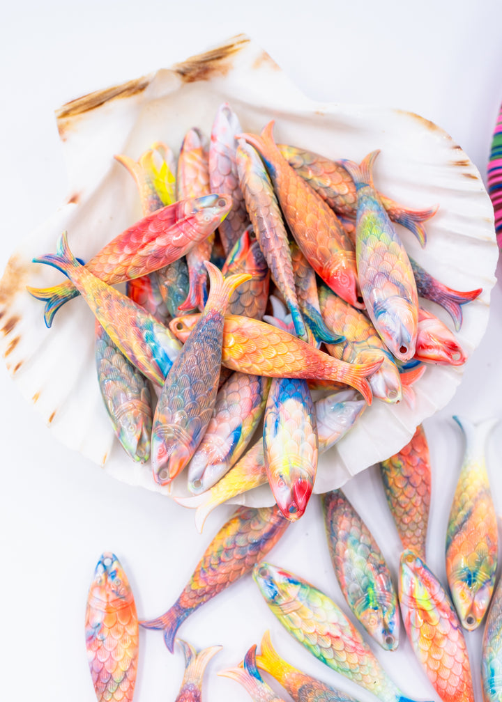 A pile of small decorative fish-shaped charms made from German resin with colorful painted scales, arranged on a white shell and scattered on a white surface.