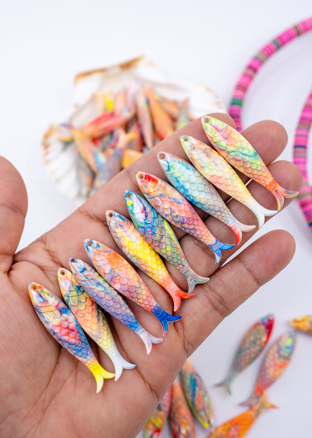 A hand holding a row of small, brightly colored German resin fish-shaped items with painted scales, with more similar items blurred in the background.
