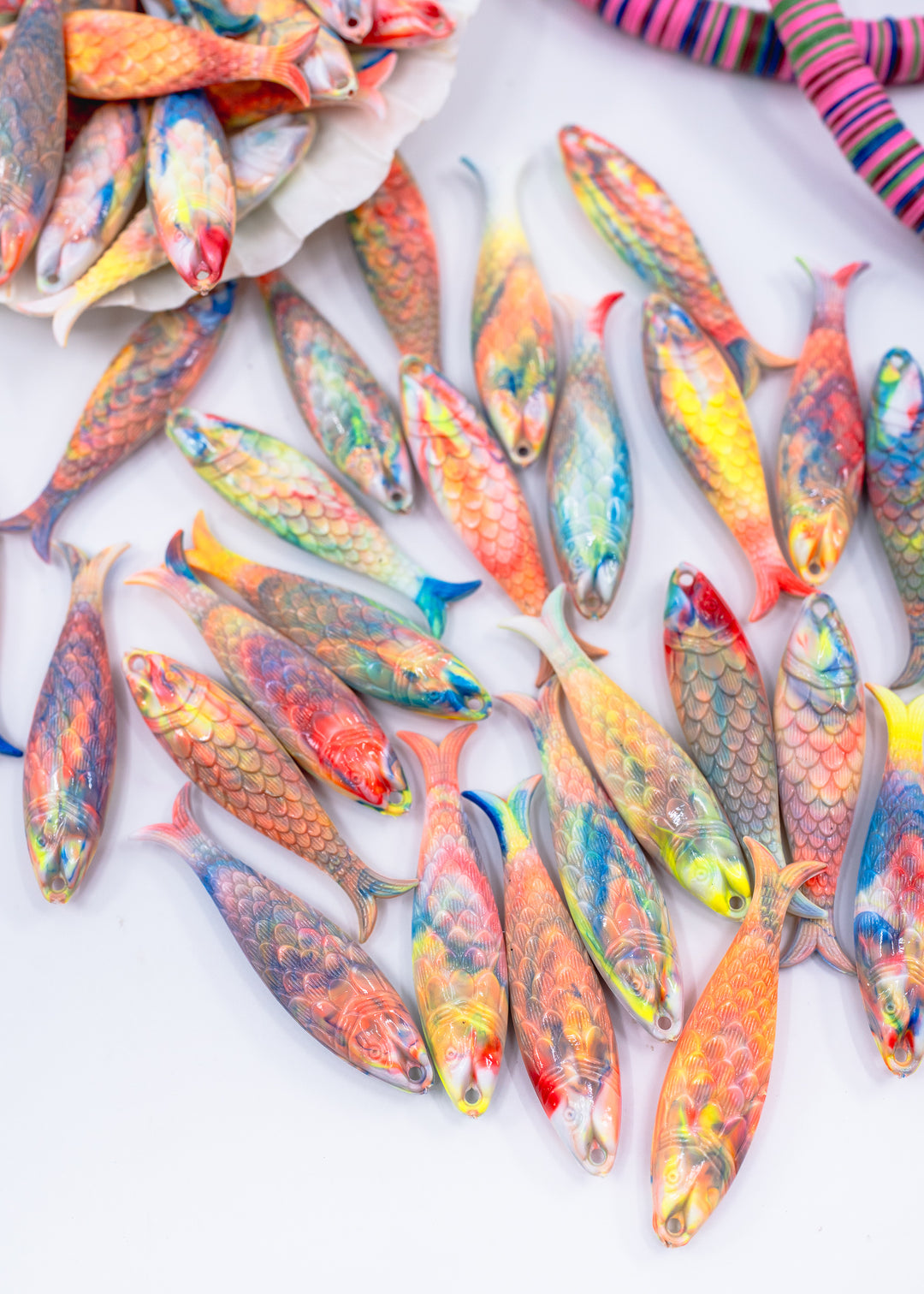 A collection of colorful, fish-shaped German resin charms with detailed scales and painted patterns, arranged on a white background with some in a shell and others scattered freely.