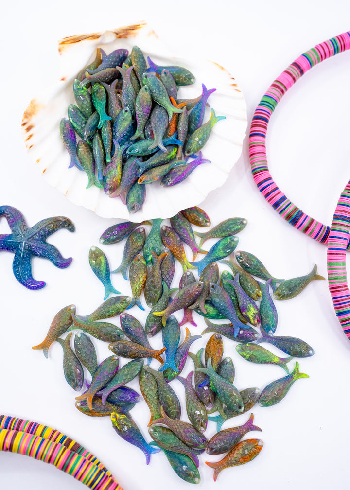 A collection of small, fish-shaped German resin charms with iridescent, multicolored scales, displayed on a white background with a seashell, alongside colorful striped jewelry cords.