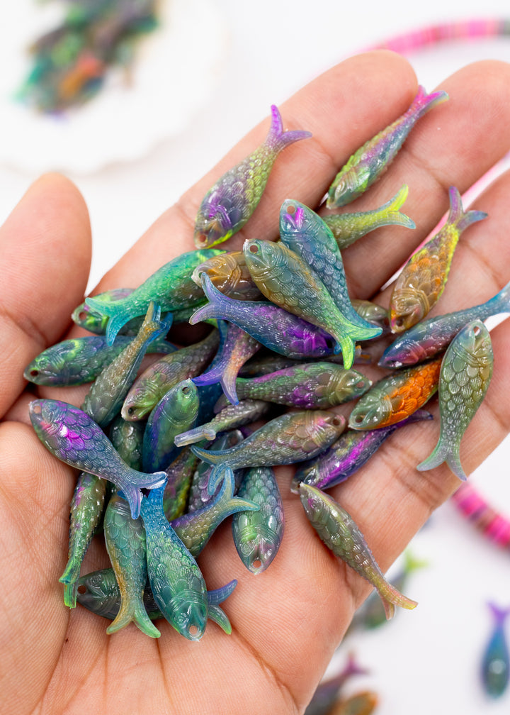 A hand holding a pile of small, iridescent fish-shaped beads made from German resin fish charms with shimmering green, blue, purple, and orange hues on a white background.