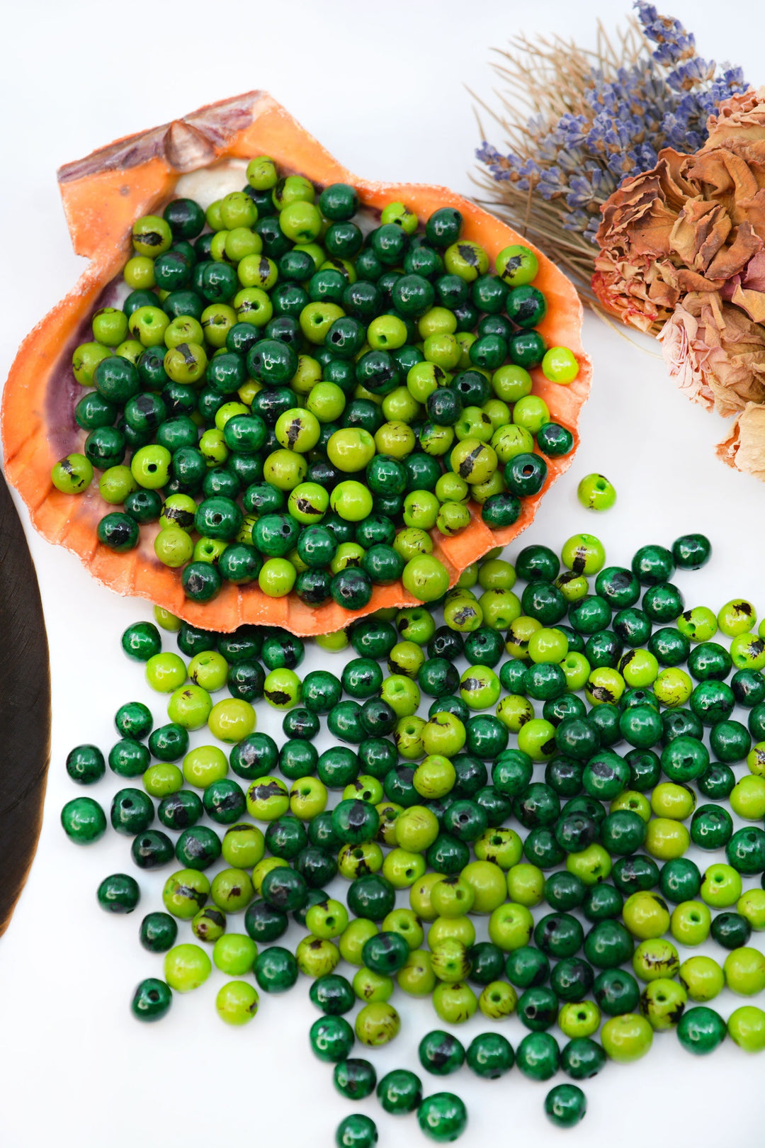 A collection of round green acai beads of varying sizes, some with a black line pattern, displayed on a white surface with an orange fabric and a sprig of lavender in the background.