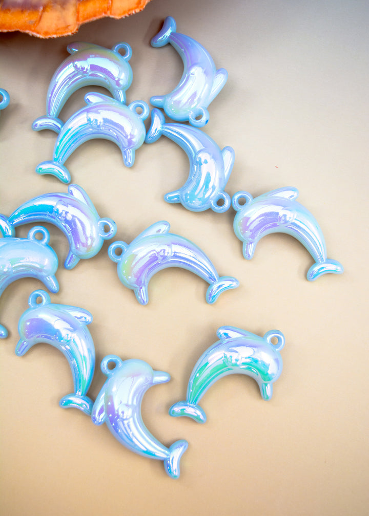 Several small dolphin-shaped charms with a glossy iridescent blue and lavender finish scattered on a beige surface.