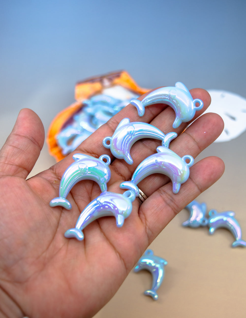 A hand holding several small iridescent dolphin-shaped charms or pendants, with more of the same visible in the background on a beige surface against a soft blue backdrop.