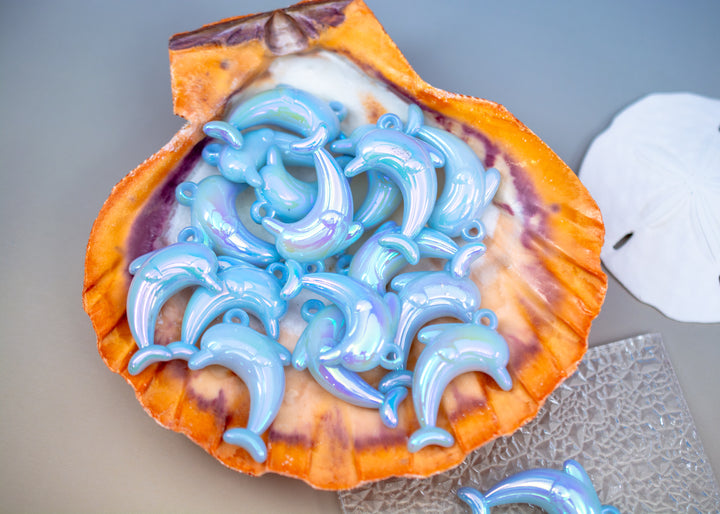 A collection of glossy, iridescent light blue dolphin pendants arranged inside an open orange and purple seashell, with a white sand dollar and a textured clear plate on a gray surface.