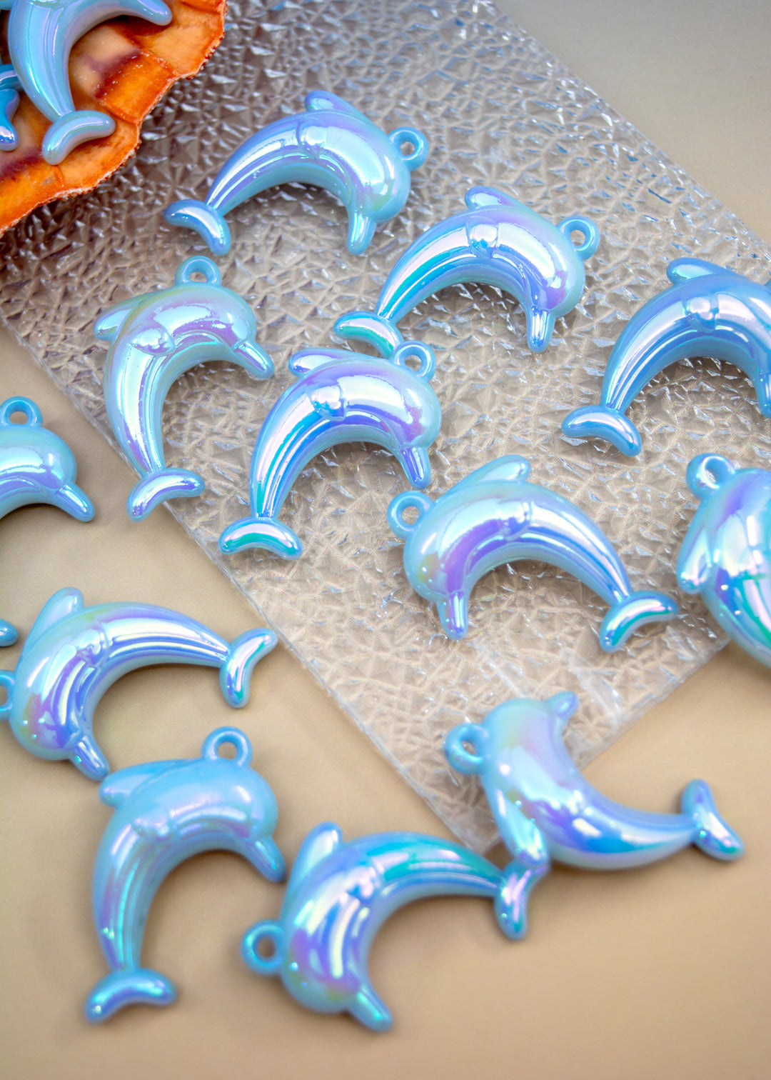 Several iridescent dolphin-shaped jewelry charms or pendants arranged on a textured clear sheet over a beige surface.