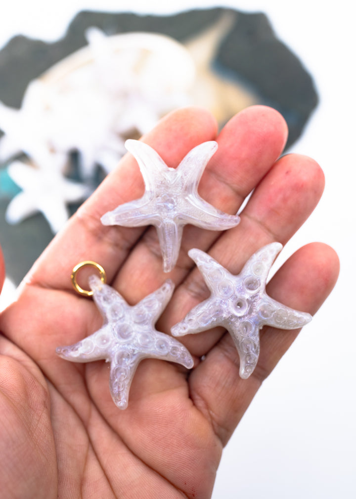 Three small translucent lavender starfish charms made from German resin with gold loops, held in a person&