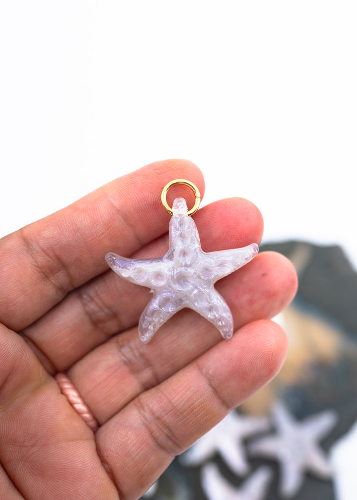 A hand holding a small translucent lavender starfish pendant charm made from German resin with a gold ring clasp, against a bright white background with a blurred dark object below.