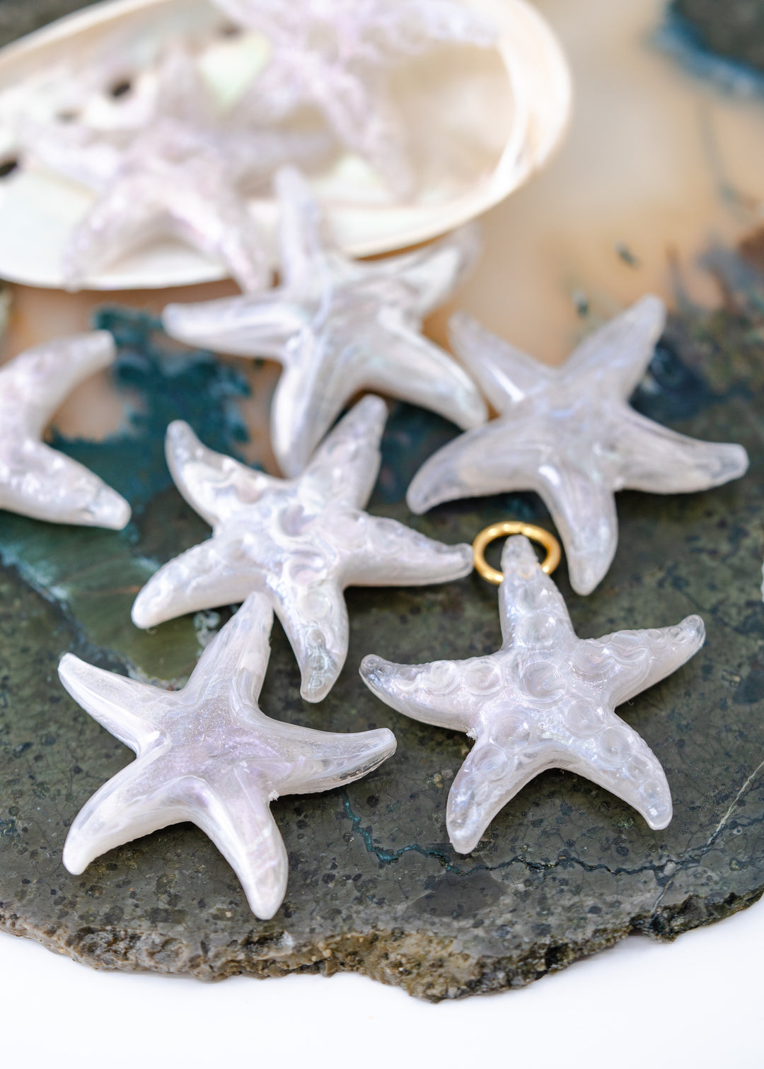 Several translucent white starfish-shaped jewelry charms made from German resin with small gold loops are arranged on a dark stone surface, with a shell visible in the background.