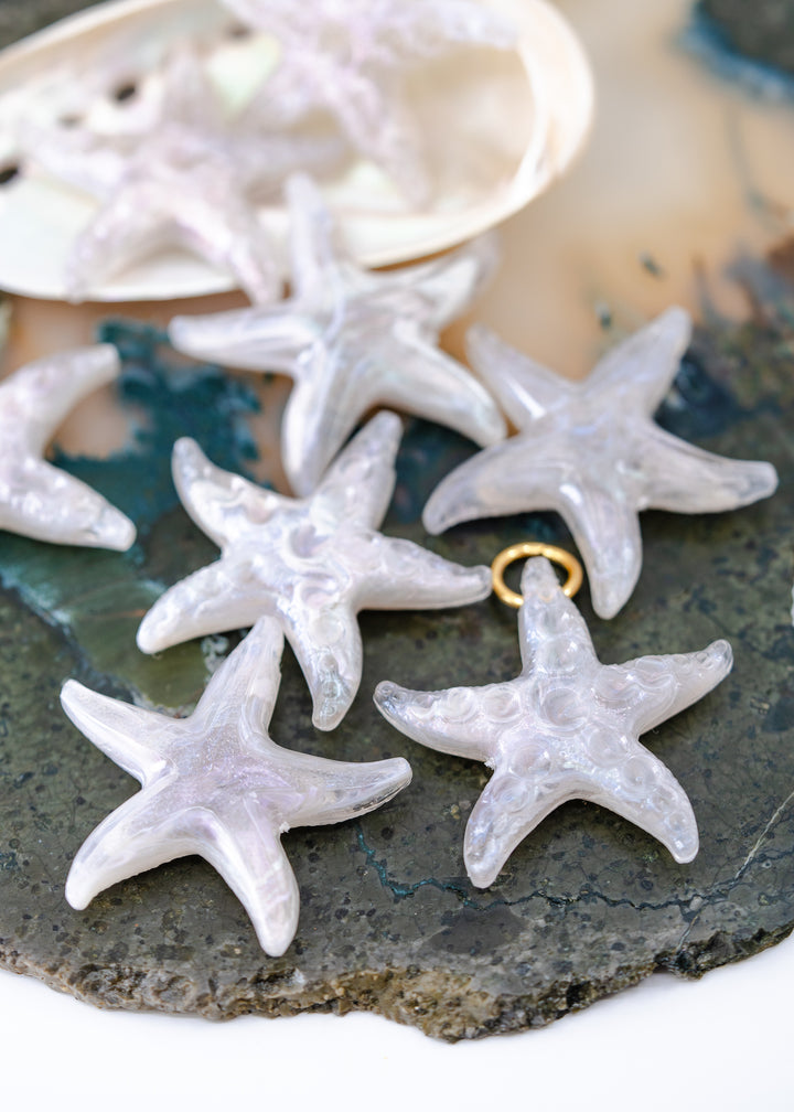Several translucent white starfish-shaped jewelry charms made from German resin with small gold loops are arranged on a dark stone surface, with a shell visible in the background.