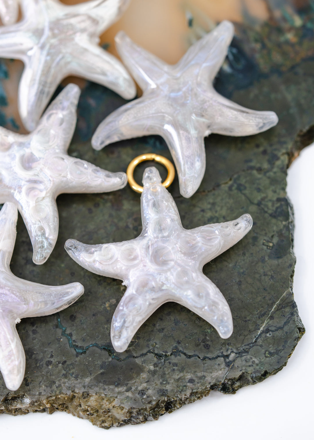 Clear iridescent starfish-shaped jewelry pendants made from German resin with a small gold jump ring, displayed on a dark textured stone surface.