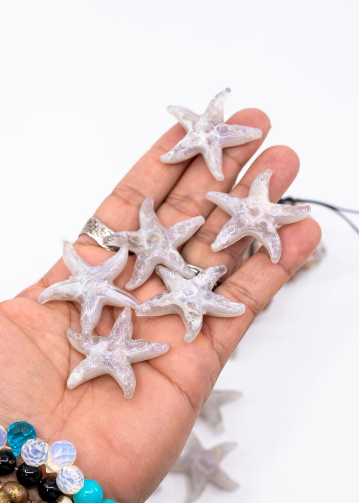 A hand holding several translucent white starfish-shaped jewelry charms made from German resin or beads against a plain white background.