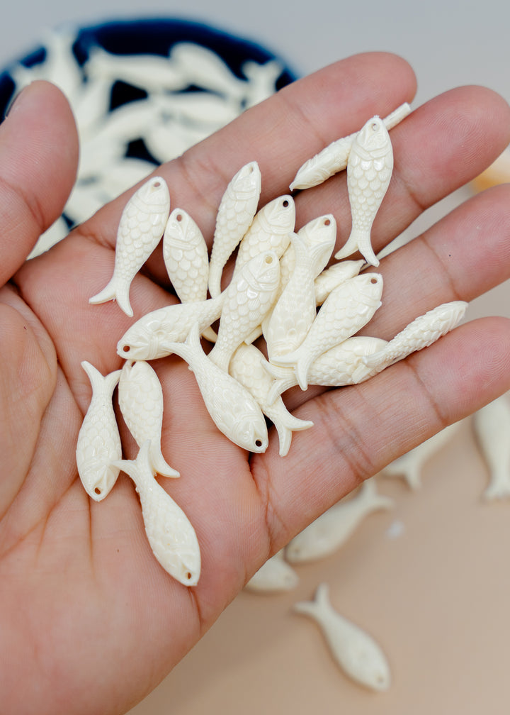 A hand holding several small white fish-shaped beads or charms with textured scales.