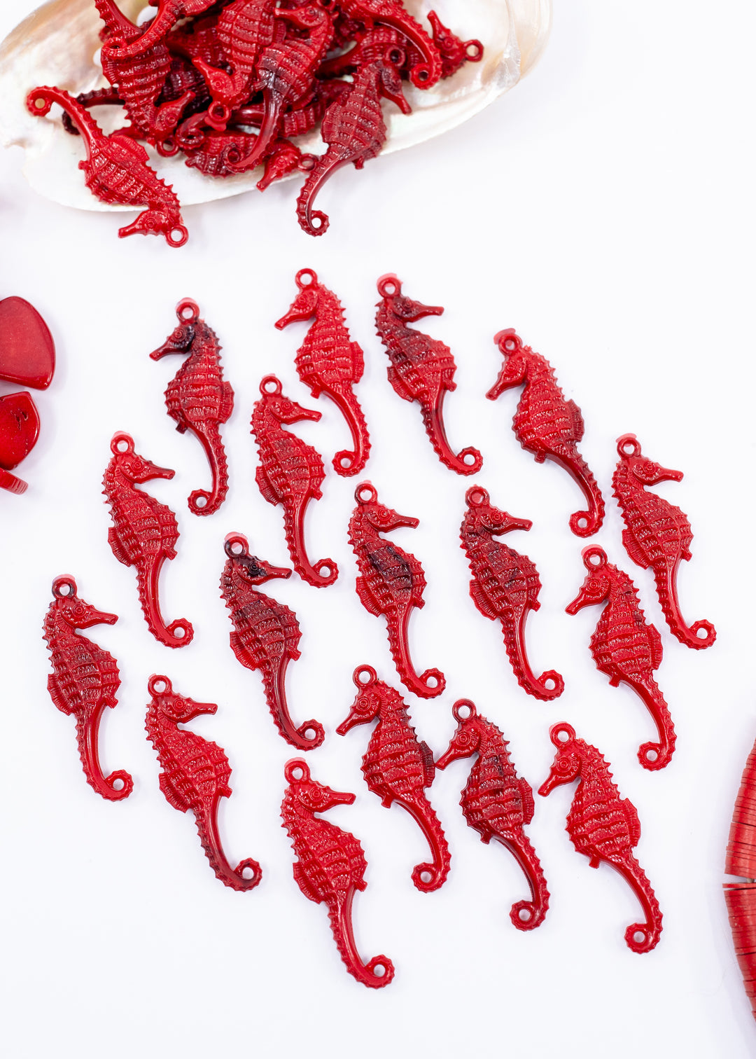 Multiple red seahorse-shaped jewelry charms or beads arranged on a white background, with a small pile of similar pieces visible at the top and a partial red object at the lower right edge.