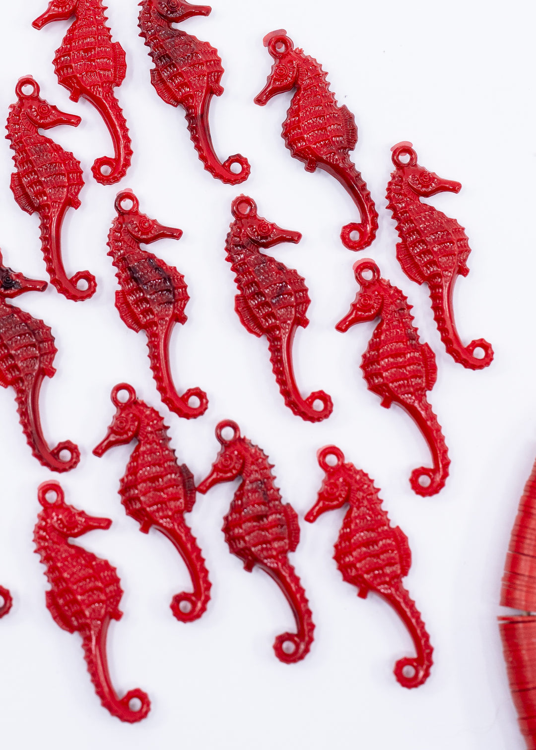Multiple small red seahorse-shaped jewelry charms made from German resin scattered on a plain white background, with a partial matching red object visible at the lower right edge.