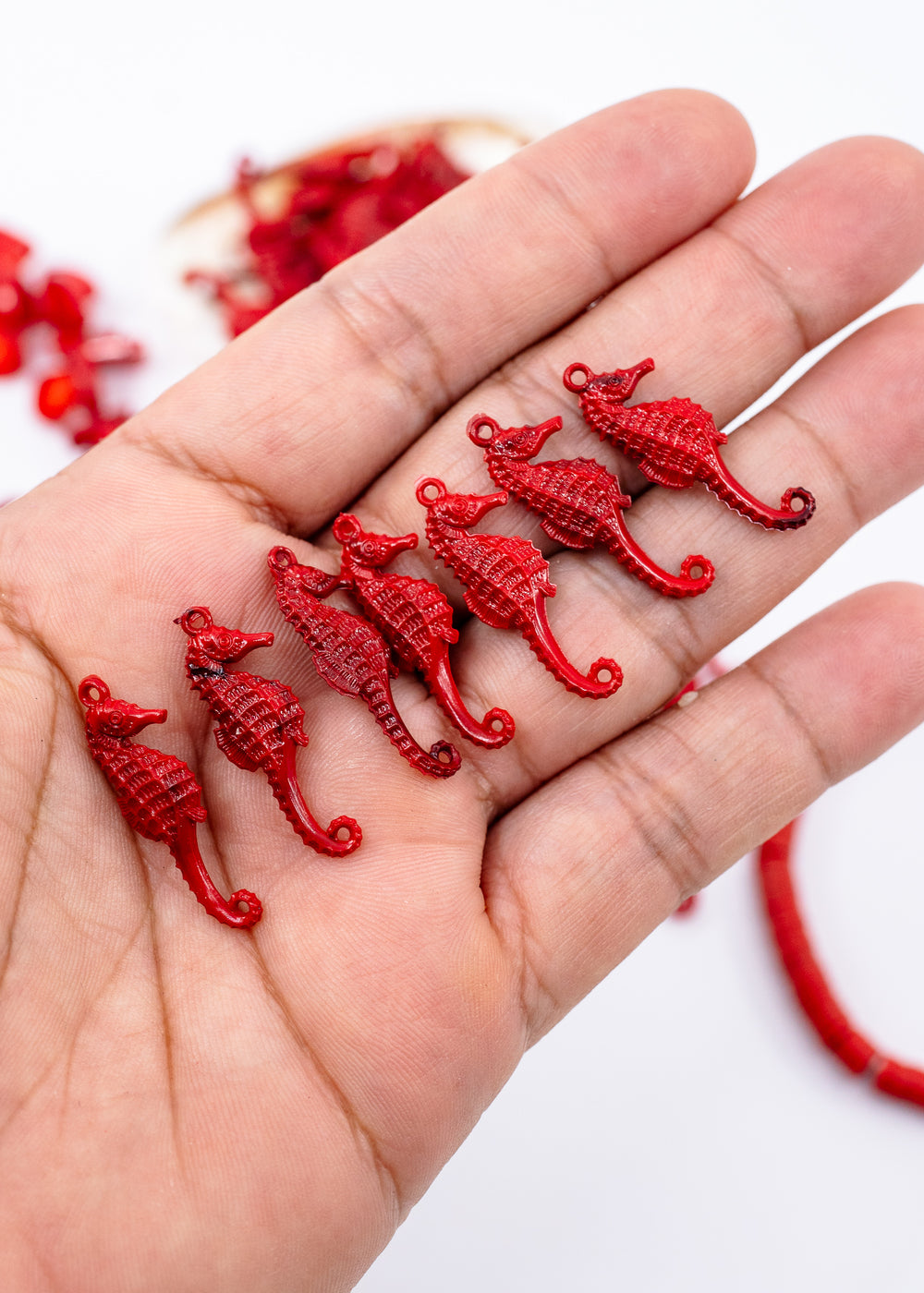 A hand holding several small red seahorse-shaped beads or charms, made from German resin with a few more red beads blurred in the background on a white surface.