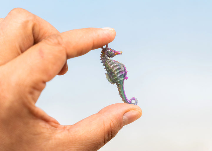 A hand holding a small seahorse-shaped pendant or charm made from German resin against a soft blue sky background.