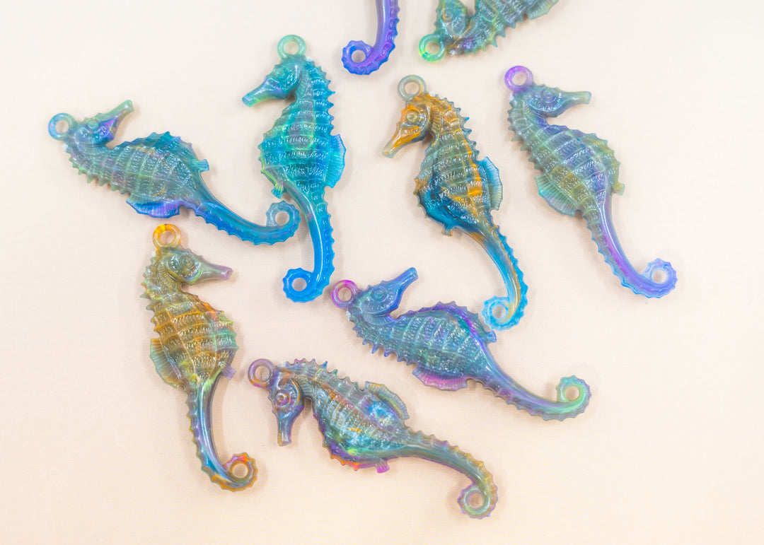 A set of iridescent seahorse-shaped jewelry charms or pendants scattered on a plain off-white background. German resin charms for jewelry making.