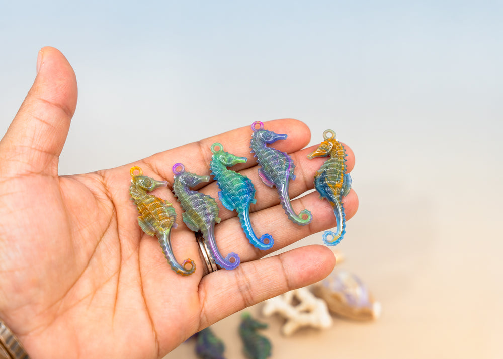 Five small iridescent German Resin seahorse charms resting on an open hand, with a soft blurred beach-like background.