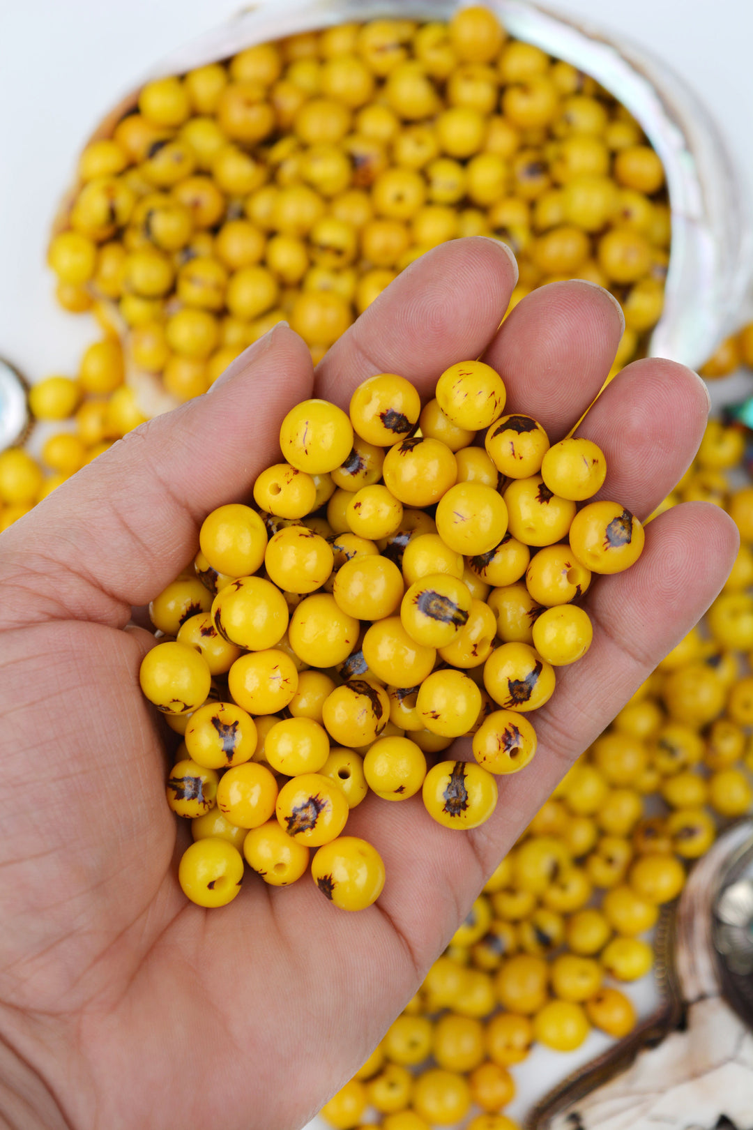 A hand holding a small container filled with sunny yellow acai beads, with another container full of beads visible in the background.