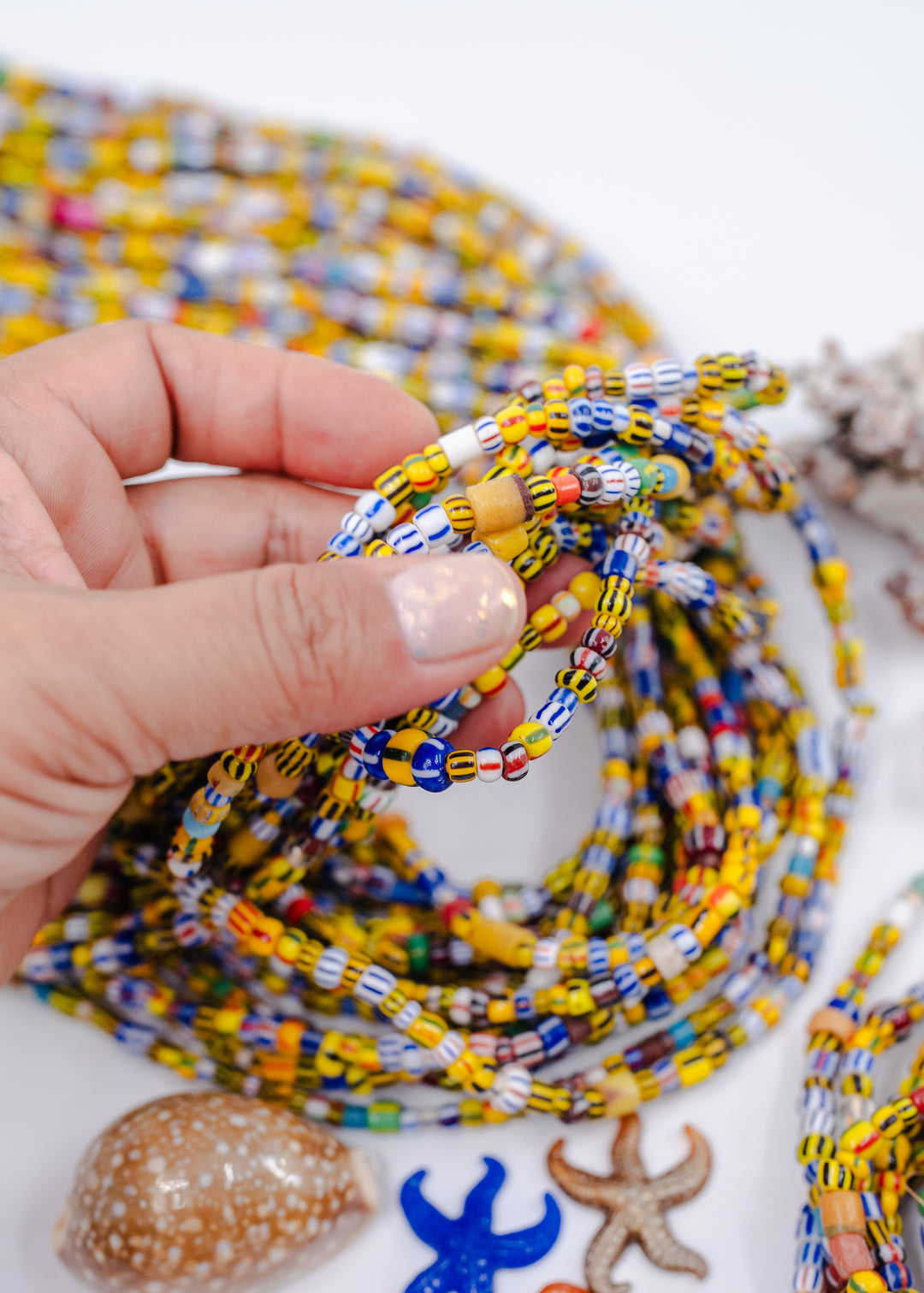 A close-up of a hand holding a coiled strand of colorful African Glass Beads, in stripped yellow and other colors. For necklace or bracelet, with yellow, blue, white, and orange beads. Seashells and starfish decorations are arranged on a white surface in the foreground.