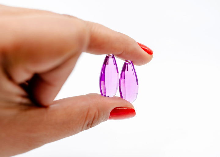 A close-up of a hand holding a faceted purple teardrop-shaped German resin bead.