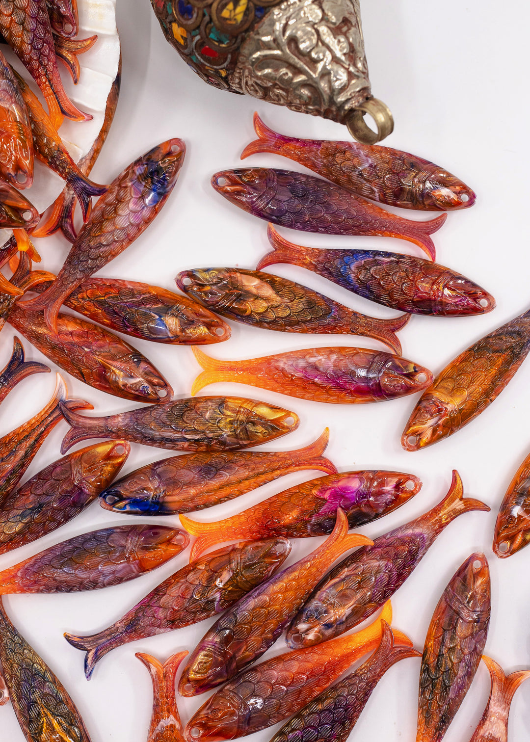 A collection of small, handcrafted fish-shaped charms with detailed scales and vibrant orange, red, and brown colors, arranged on a white surface with an ornate metal container partially visible at the top. Made from German resin.