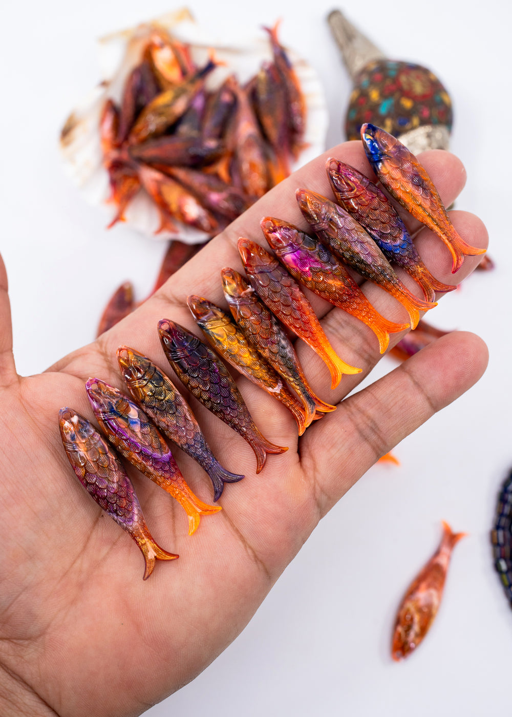 A hand holds a row of small, intricately patterned fish-shaped charms in various vibrant colors like orange, purple, blue, and gold, with more of the same and a larger matching object blurred in the background on a white surface.