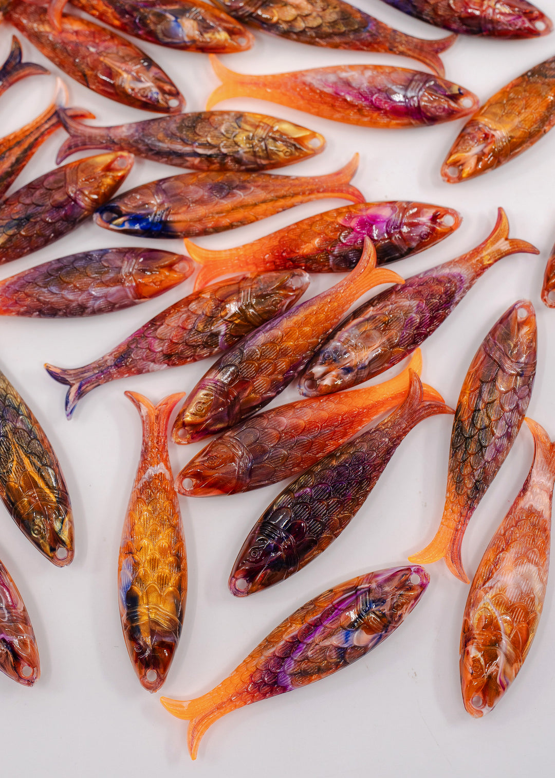 A collection of small, handcrafted resin fish charms with detailed scales and vibrant orange, purple, and brown colors, arranged on a plain white background. Made in Germany.