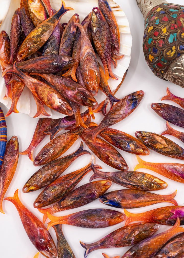A close-up overhead view of many small decorative fish charms made from German resin arranged on a white surface, with some clustered in a shell and one ornate multicolored object visible at the upper right.