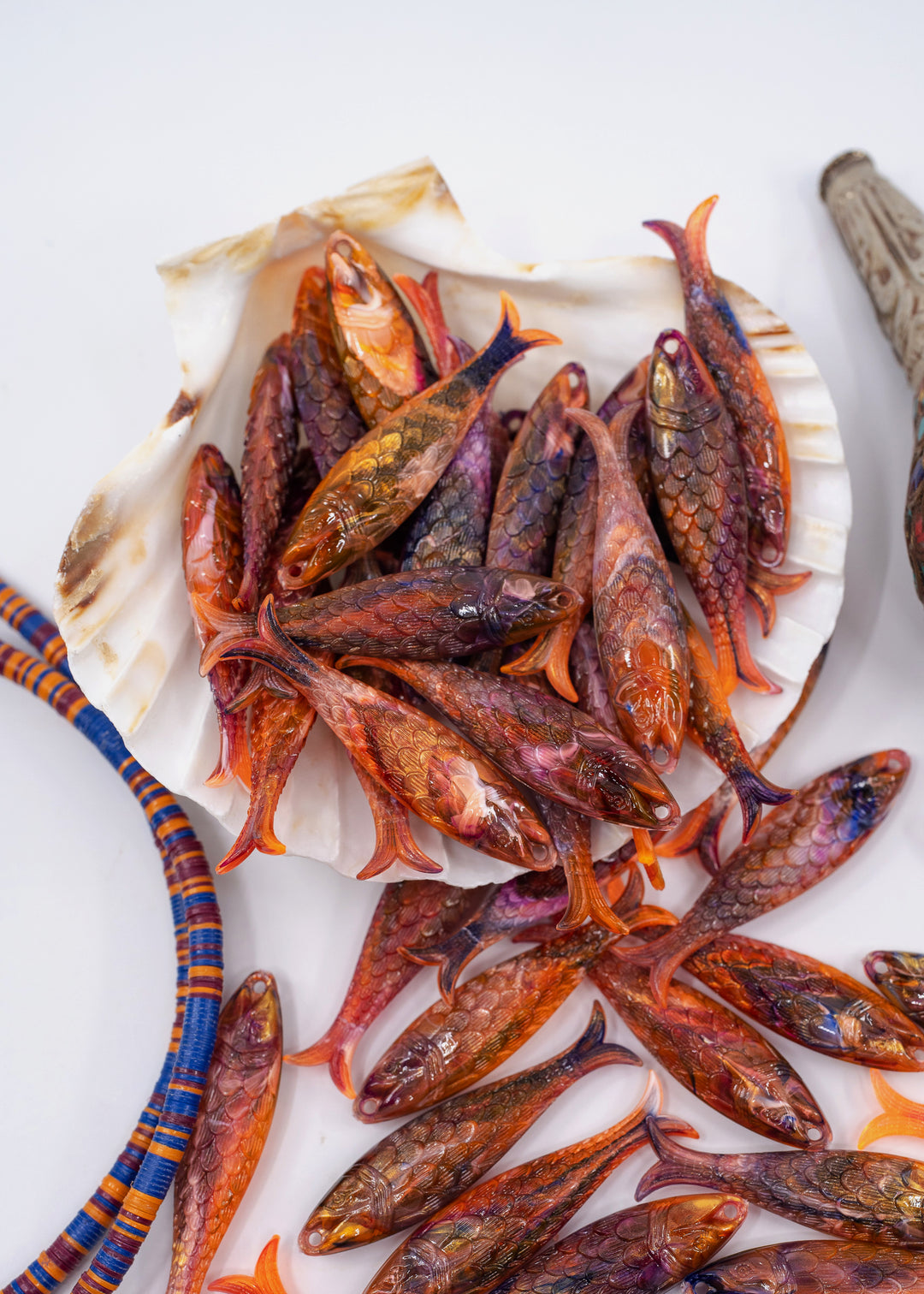 Small decorative fish-shaped charms or beads in orange, red, and brown tones, arranged on a white shell and scattered on a white surface, with partial colorful woven cords visible at the edges. Made in Germany from recycled resin.